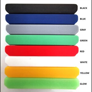 May include: A stack of rectangular, solid objects in various colors: black, blue, gray, green, red, white, yellow, and glow-in-the-dark green. Each object has a rounded edge and a textured surface. The colors are labeled with corresponding text.
