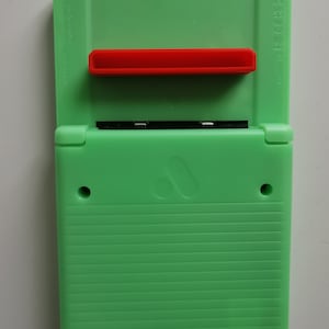 May include: A bright green handheld gaming device with a red button and a black screen. The device has a ribbed texture on the bottom half and a smooth top. The device has a logo on the front.