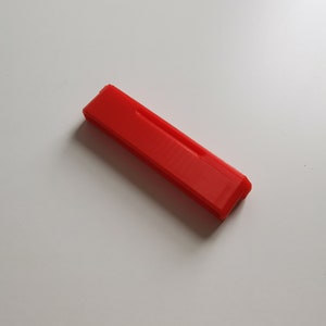 May include: A bright red, rectangular object with a slightly textured surface. The object has a beveled edge on one end and a recessed groove along its length. It is lying on a white surface.