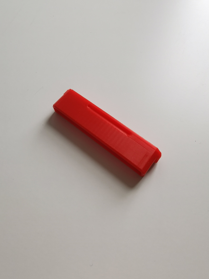 May include: A bright red, rectangular object with a slightly textured surface. The object has a beveled edge on one end and a recessed groove along its length. It is lying on a white surface.
