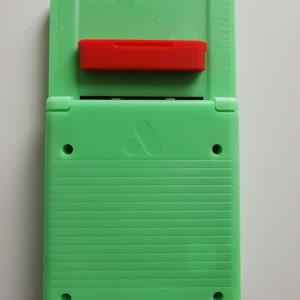 May include: A mint green handheld gaming device with a red cartridge inserted. The device is open, revealing the screen area. The back has horizontal ridges and two small circular holes. The device has a logo on the bottom half.