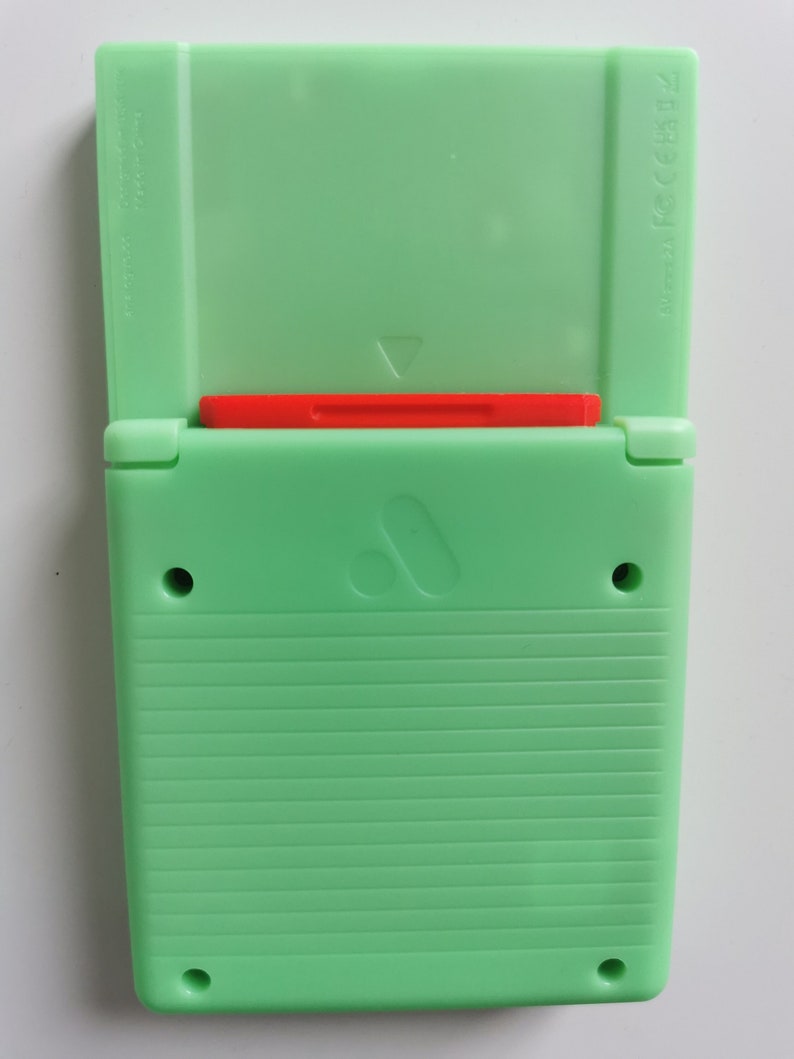 May include: A mint green handheld gaming device with a red cartridge slot. The device is open, revealing the interior. The back has horizontal ridges and two circular holes. A white surface provides the background.