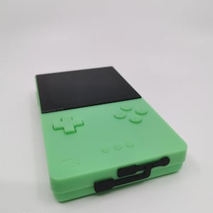 May include: A mint green handheld gaming device with a black top section and black buttons. The device has a cross-shaped directional pad and four circular buttons. The text "ADD ON: Connection port dust cover (soft plastic, only black)" is visible.