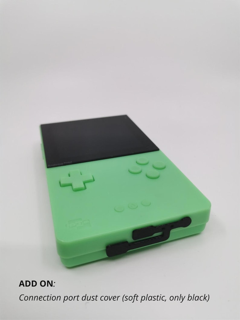 May include: A mint green handheld gaming device with a black top section and black buttons. The device has a cross-shaped directional pad and four circular buttons. The text "ADD ON: Connection port dust cover (soft plastic, only black)" is visible.