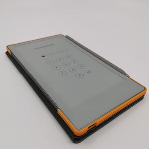 May include: A gray tablet with an orange frame and a stylus, resting on a black case. The screen displays a numeric keypad with the text "Enter passcode" at the top. The stylus is gray.