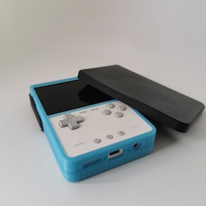 May include: A handheld gaming console with a blue and white case, featuring a black screen and gray buttons. A black cover rests on top. The device has a directional pad and several control buttons.