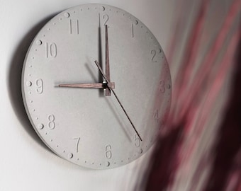 Può includere: Un orologio da parete rotondo, grigio chiaro, con numeri in rilievo e piccoli punti sul bordo. Le lancette dell'orologio sono in legno scuro. L'ora indicata è circa le 9:05. L'orologio è montato su una parete bianca.