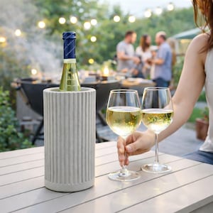 Könnte beinhalten: Ein grauer, geriffelter Weinkühler hält eine Flasche Weißwein. Zwei Weingläser, gefüllt mit Weißwein, werden vor dem Kühler gehalten. Die Szene spielt sich im Freien mit Lichterketten und Personen im Hintergrund ab.