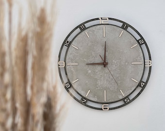Può includere: Un orologio rotondo grigio con numeri neri e una lancetta marrone. L'orologio ha un quadrante testurizzato, simile al cemento.