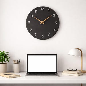 Könnte beinhalten: Eine schwarze runde Wanduhr mit weißen Ziffern und hellbraunen Zeigern hängt an der Wand. Ein Laptop, Bücher, eine Lampe und eine Topfpflanze stehen auf dem weißen Schreibtisch. Der Durchmesser der Uhr beträgt ca. 30 cm.