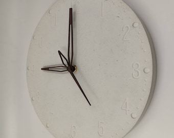 Può includere: Un orologio da parete rotondo, bianco sporco, con numeri in rilievo e una superficie strutturata. Le lancette dell'orologio sono marrone scuro e segnano circa le 9:16. L'orologio ha un design minimalista.
