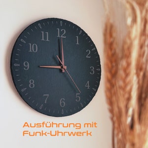Op de afbeelding: Een zwarte leisteen klok met witte cijfers en een koperkleurige uur- en minutenwijzer. De klok is gemonteerd op een witte muur. De tekst "Ausführung mit Funk-Uhrwerk" is onder de klok gedrukt.