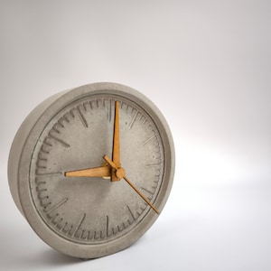 Könnte beinhalten: Eine graue Betonuhr mit goldfarbenen Zeigern. Das Zifferblatt der Uhr hat ein schlichtes Design mit schwarzen Markierungen für Stunden und Minuten.