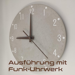 Può includere: Un orologio rotondo in cemento bianco con lancette nere e numeri bianchi. L'orologio ha uno sfondo bianco con una texture sottile. Il quadrante dell'orologio indica "Ausführung mit Funk-Uhrwerk".