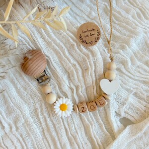 Op de afbeelding: Houten fopspeenclip met de naam "ZOE" gespeld in houten letterkralen. De clip heeft een witte madelief, een wit hart en houten kralen. Een klein, rond label met "handmade with love" is ook aanwezig.