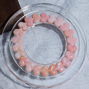 Puede incluir: Una pulsera circular compuesta por cuentas lisas en forma de corazón de color rosa claro dispuestas en un bucle continuo. Las cuentas tienen un acabado brillante y pulido, y son uniformes en tamaño y forma, creando un patrón consistente.