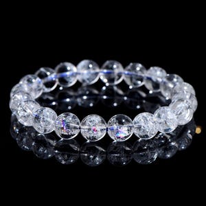 May include: A clear quartz crystal bead bracelet. The round beads are transparent with internal fractures and iridescent inclusions. The bracelet is strung on a blue elastic cord, set against a black background.