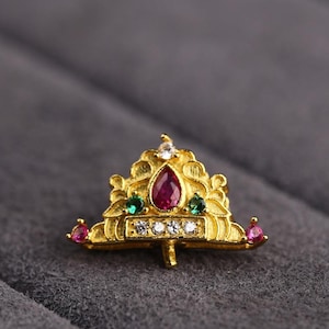 May include: A gold-colored crown-shaped jewelry piece with a central teardrop-shaped ruby, surrounded by smaller green and white gemstones. The crown has a detailed, ornate design and is set against a gray background.
