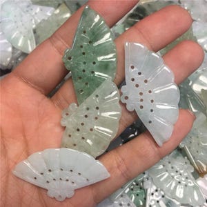 Pode incluir: Uma coleção de pingentes em forma de leque de jade verde e branco com intrincados desenhos recortados. Os pingentes são feitos de um material translúcido e têm uma estética delicada e feminina.