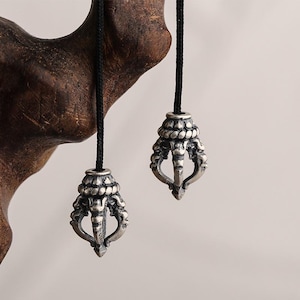 Könnte beinhalten: Zwei silberfarbene Anhänger mit aufwendigen Designs hängen an schwarzen Kordeln. Die Anhänger haben eine detaillierte, verzierte Form, möglicherweise ein religiöses oder kulturelles Symbol. Der Hintergrund ist unscharf und neutral.