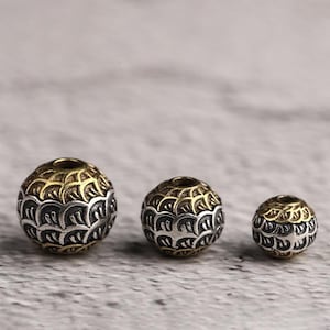 Op de afbeelding: Drie decoratieve metalen kralen met een gedetailleerd patroon. De kralen zijn bolvormig, met een zilver- en goudkleurenschema. Het ontwerp heeft een herhalend, bladachtig patroon. De kralen variëren in grootte.