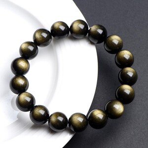 K&ouml;nnte beinhalten: Ein Armband aus schwarzen und goldenen Perlen mit gl&auml;nzendem, irisierendem Finish. Die Perlen sind rund und haben eine glatte Oberfl&auml;che.