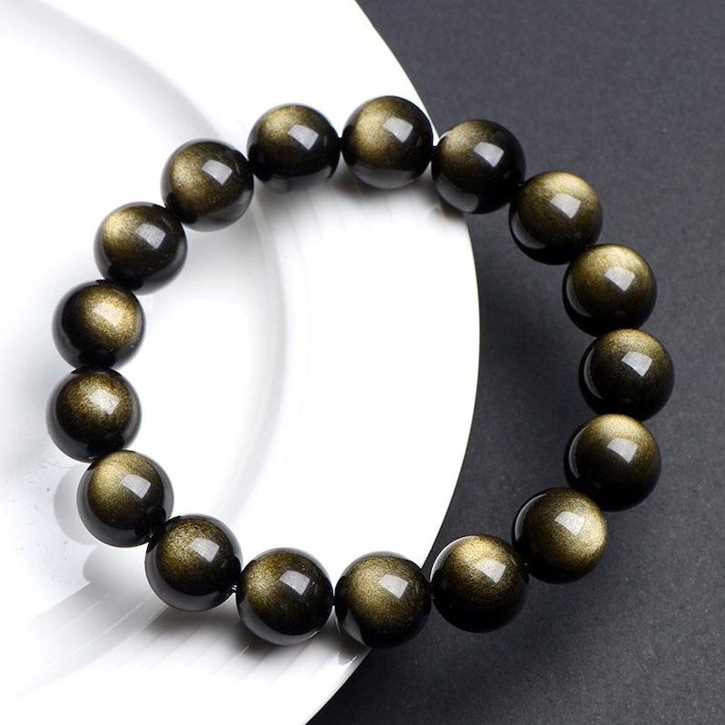 K&ouml;nnte beinhalten: Ein Armband aus schwarzen und goldenen Perlen mit gl&auml;nzendem, irisierendem Finish. Die Perlen sind rund und haben eine glatte Oberfl&auml;che.