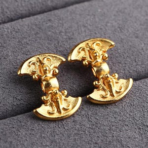 May include: A pair of gold-colored earrings. Each earring features a stylized design with a crescent shape at the top and bottom, connected by a central element. The earrings have an ornate, detailed pattern.
