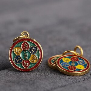 Puede incluir: Dos colgantes de color dorado con un dise&ntilde;o circular. Los colgantes est&aacute;n decorados con un patr&oacute;n de mandala colorido con cinco p&eacute;talos en rojo, amarillo, azul y verde. Cada p&eacute;talo tiene un s&iacute;mbolo diferente escrito en negro.