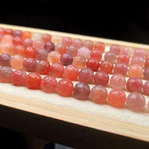 Könnte beinhalten: Reihenweise geschnitzte, runde Perlen in Rot-, Orange- und Lilatönen, die in einem Holztablett präsentiert werden. Die Perlen weisen ein detailliertes, blumenartiges Muster auf und sind auf einem cremefarbenen Stoff angeordnet.