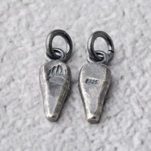 May include: Two silver-toned pendant charms with a teardrop shape and a small loop at the top. One charm has a bear paw print, the other has a small inscription. The charms are on a light gray background.