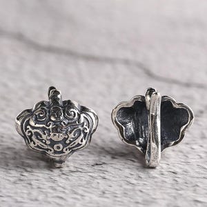 May include: Two silver-colored, ornate pendants. Each pendant has a detailed, symmetrical design with swirling patterns and a central motif. One pendant is viewed from the front, the other from the side, showing a loop for hanging.