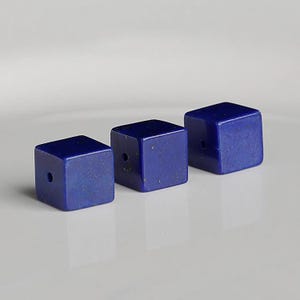 Puede incluir: Tres cuentas cúbicas de color azul oscuro, cada una con un agujero perforado. Las cuentas están dispuestas en fila, cada una con un ángulo ligeramente diferente. Las cuentas tienen una superficie lisa y pulida.