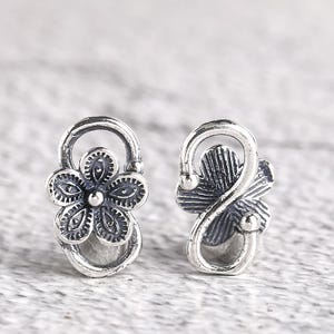 May include: A pair of silver-coloured stud earrings. One earring features a detailed flower design with five petals and a central bead. The other earring has a stylized leaf design. Both earrings have a curved top and a loop.