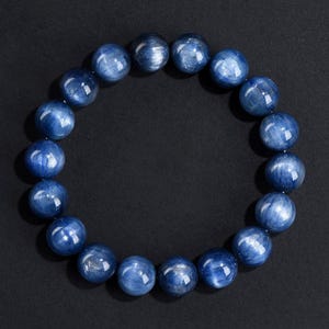 Könnte beinhalten: Ein Armband aus runden, polierten blauen Kyanit-Perlen. Die Perlen haben eine glänzende Oberfläche und ein wirbelndes, faseriges Aussehen. Das Armband wird vor einem dunklen Hintergrund präsentiert, der die leuchtende blaue Farbe und die natürlichen Muster der Steine hervorhebt.