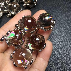 以下が含まれることがあります： 銀色の金属フレームにセットされた、球形で半透明の茶色のクリスタルペンダント。各球体は、虹色の光の屈折を内部に示しています。滑らかで磨かれた石は丸みを帯びており、装飾的な台座に取り付けられています。