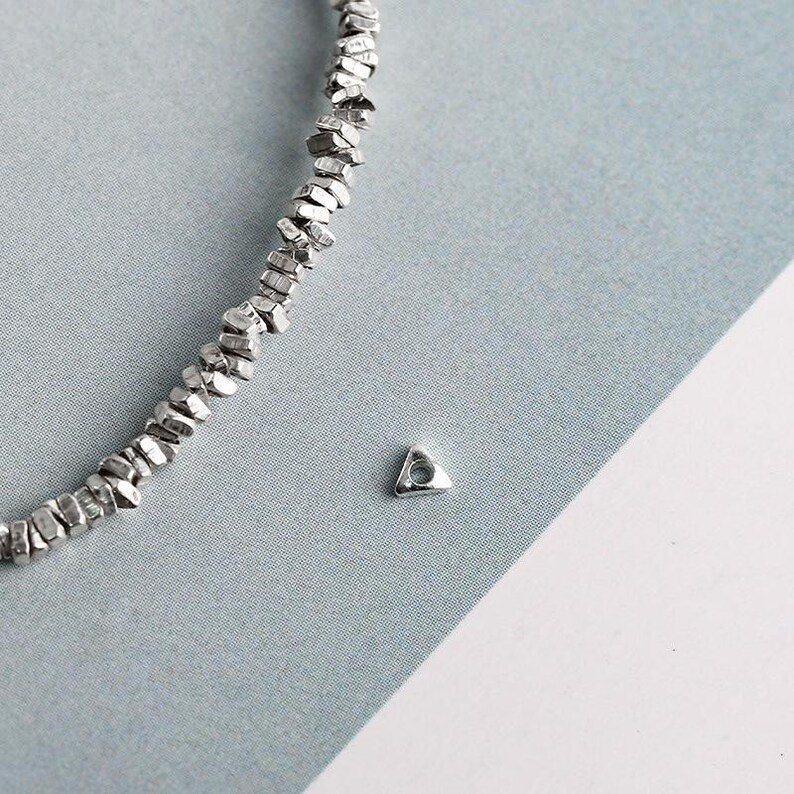 May include: A silver-toned necklace with a textured, irregular bead design. A small, triangular charm with a hole is also visible. The jewelry is displayed on a light blue and white background.