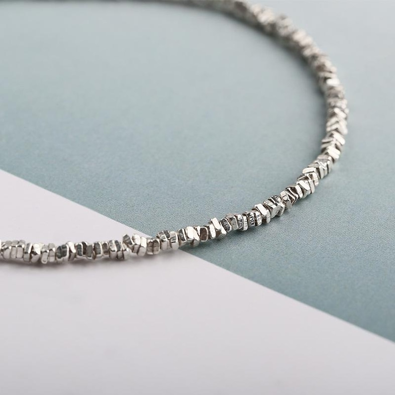May include: A delicate silver necklace featuring small, irregularly shaped beads. The beads are strung together, creating a subtle, textured appearance. The necklace is displayed against a soft, two-tone background of white and light blue.