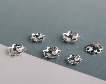 May include: Six silver-toned metal bead caps with a flower-like design. The caps are of varying sizes and are arranged on a light blue and white surface.