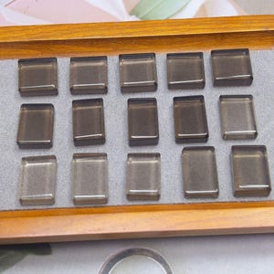 May include: A collection of fourteen rectangular, translucent, smoky-grey glass cabochons arranged in three rows on a grey fabric display tray. Each piece features a smooth, polished surface with bevelled edges, reflecting light across the top.