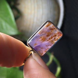 May include: A rectangular pendant with a silver-colored frame. The pendant features a polished stone with a blend of purple, orange, and brown hues. The stone's pattern resembles a natural, organic design. The pendant is held between two fingers.