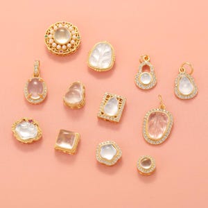 May include: A collection of gold-toned pendants and charms with various shapes, including oval, square, and teardrop. Many are adorned with clear, iridescent stones and surrounded by small, sparkling crystals. The background is a solid, soft pink.