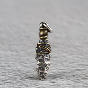 Peut inclure: Pendentif argenté en forme de pointe de flèche texturée. Il est orné d'un fil d'or et d'un enroulement en spirale. Une boucle argentée permet de l'accrocher. L'ensemble dégage une impression artisanale et rustique.