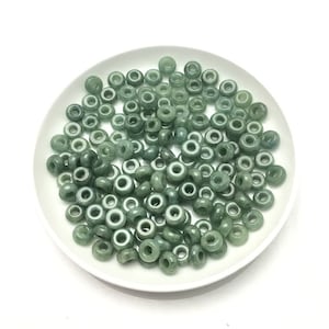 May include: A pile of green jade donut beads on a white plate. The beads are smooth and polished, with a hole in the center of each bead.