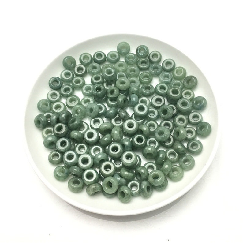 May include: A pile of green jade donut beads on a white plate. The beads are smooth and polished, with a hole in the center of each bead.