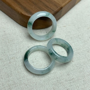 May include: Three jade rings with a translucent appearance. The rings have a light green hue with darker green veining throughout. The rings are displayed on a neutral-colored fabric and a wooden surface.