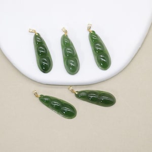 May include: Four green jade pea pod pendants with gold-coloured bails. The pendants are smooth and glossy, each with three pea-shaped segments. The pendants are arranged on a white, curved surface.