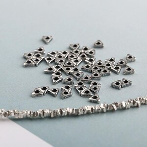 May include: A collection of small, silver-colored, triangular beads with a hole in the center, scattered on a light blue surface. A strand of similar, textured beads is visible at the bottom. These beads are suitable for jewelry making.