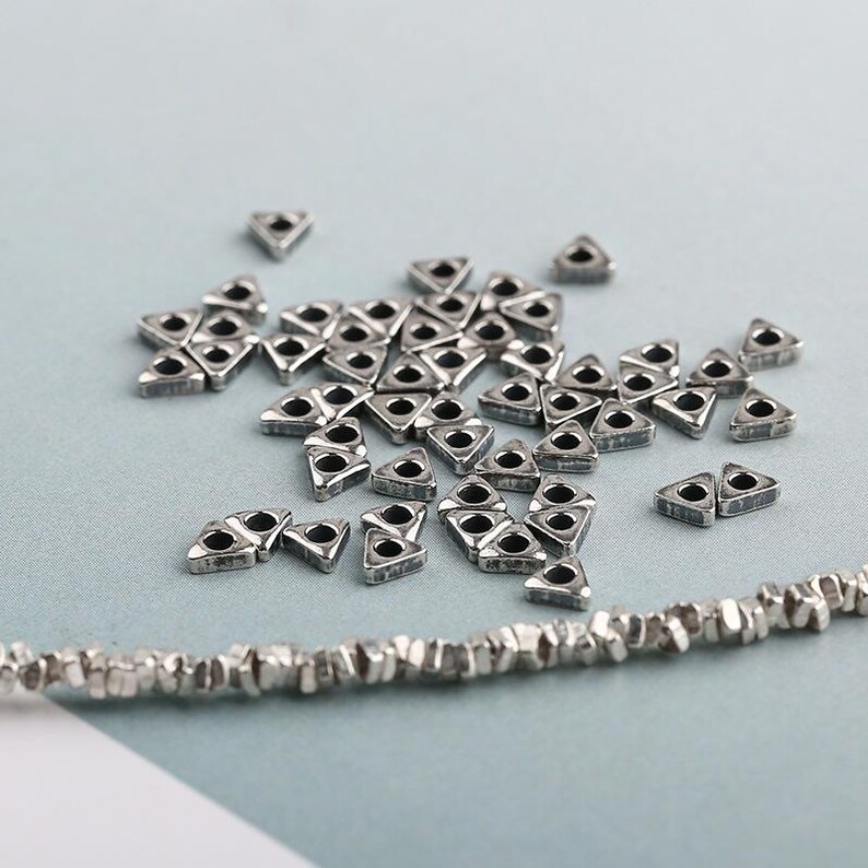 May include: A collection of small, silver-colored, triangular beads with a hole in the center, scattered on a light blue surface. A strand of similar, textured beads is visible at the bottom. These beads are suitable for jewelry making.