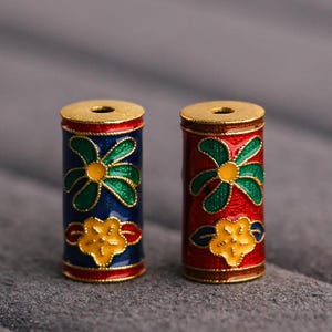 May include: Two cylindrical beads with gold trim and colourful enamel designs. The bead on the left has a blue background with green and yellow flowers. The bead on the right has a red background with green and yellow flowers.